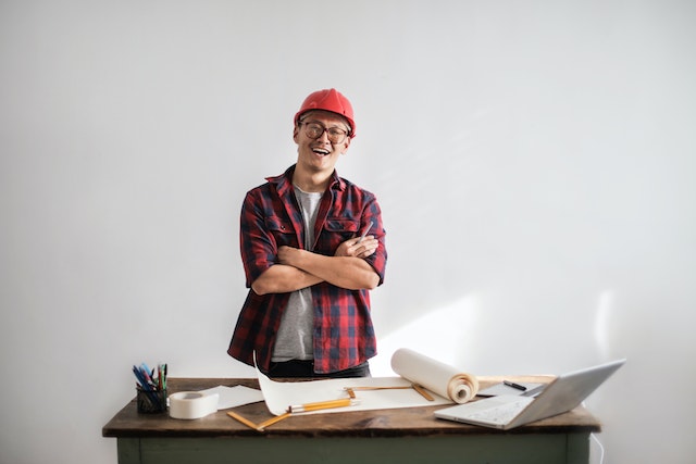 a contractor in an orange hard hat standing at desk covered in building plans